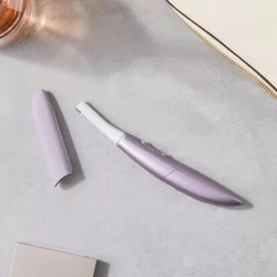 Fioletowy trymer do twarzy ze zdjętą nasadką leży na szarym blacie obok kosmetyczki i lusterka