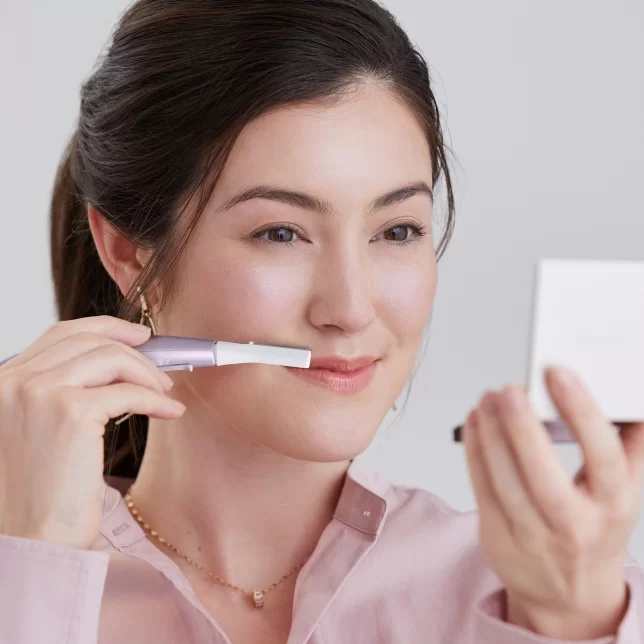 Kobieta w różowej koszuli patrzy w lusterko i używa trymera do usuwania niechcianych włosków z twarzy